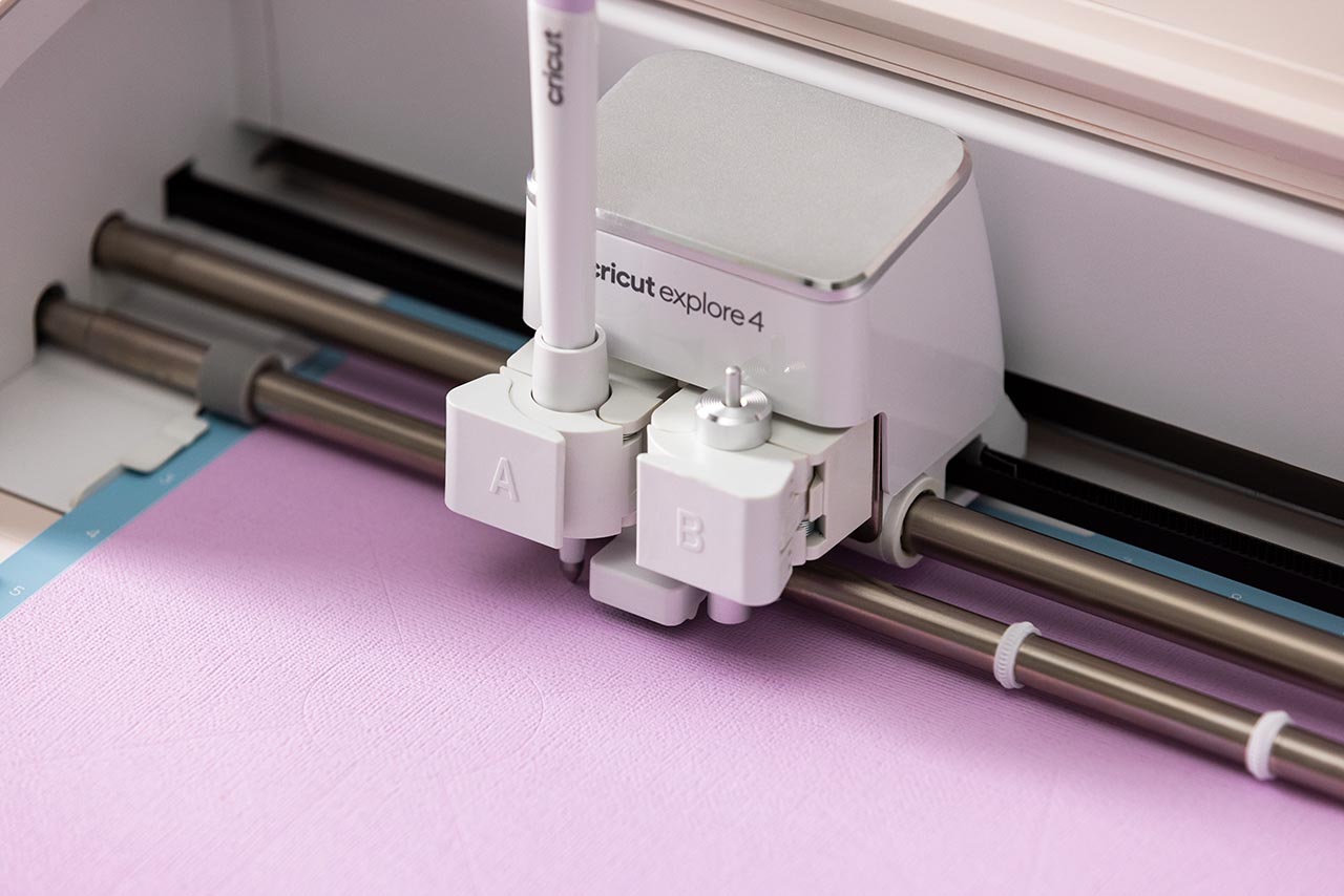 A Cricut fine-point blade housing and a scoring stylus next to heavyweight cardstock and a green StandardGrip mat on a craft table