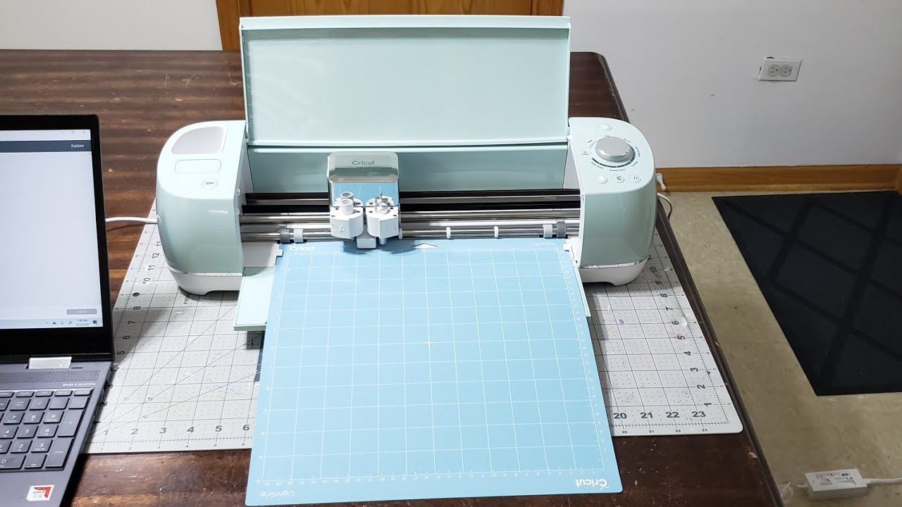 A Cricut machine with a cutting mat partially loaded into the front slot, showing the mat aligned between the guide rails on a craft table